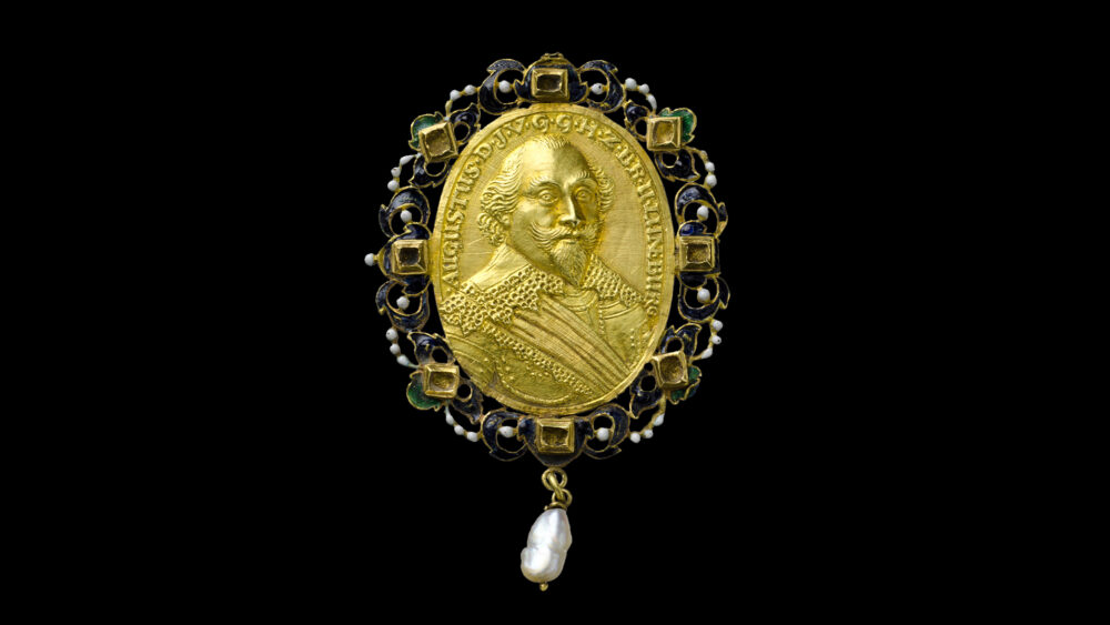 Eine goldene, ovalförmige Münze, bedruckt mit dem Portrait eines Mannes, mit filigraner Randverzierung in blau und grün und einer an der Unterseite befestigten Perle.